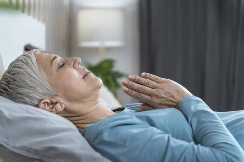 Evening meditation for promotion of sleep.  Beautiful mature woman lying in bed and meditating with hands in prayer and eyes closed.