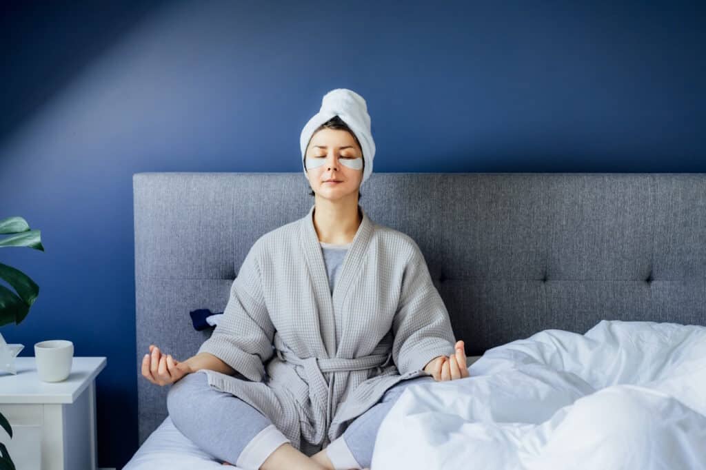 Woman in pajamas and bathrobe with eyes patches and towel on her head practicing meditation on bed