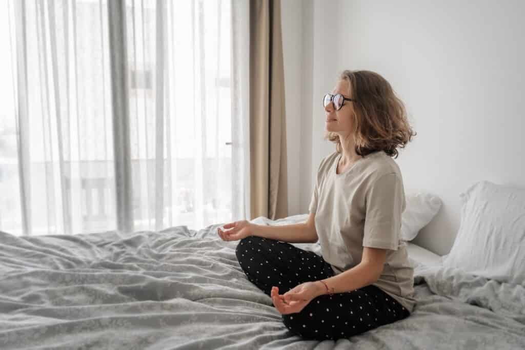 Beautiful young curly woman in glasses sits on a bed in a lotus position with her eyes closed. Mindfulness meditation and relaxation at home